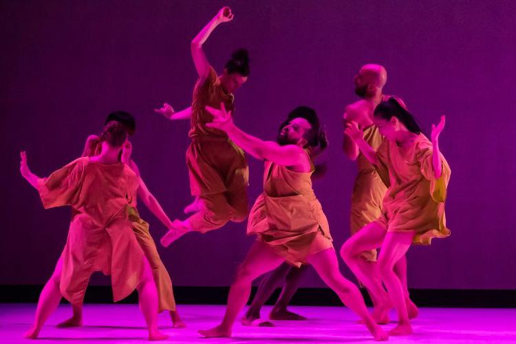 A group of dancers in mustard costumes on stage in pink/purple lighting.